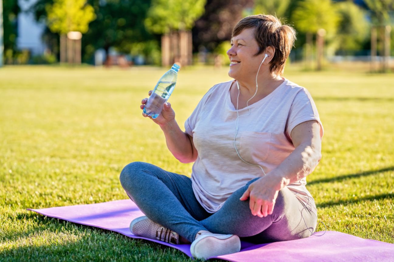 Eine Frau mittleren Alters sitzt mit Trinkflasche auf Yogamatte im Park.